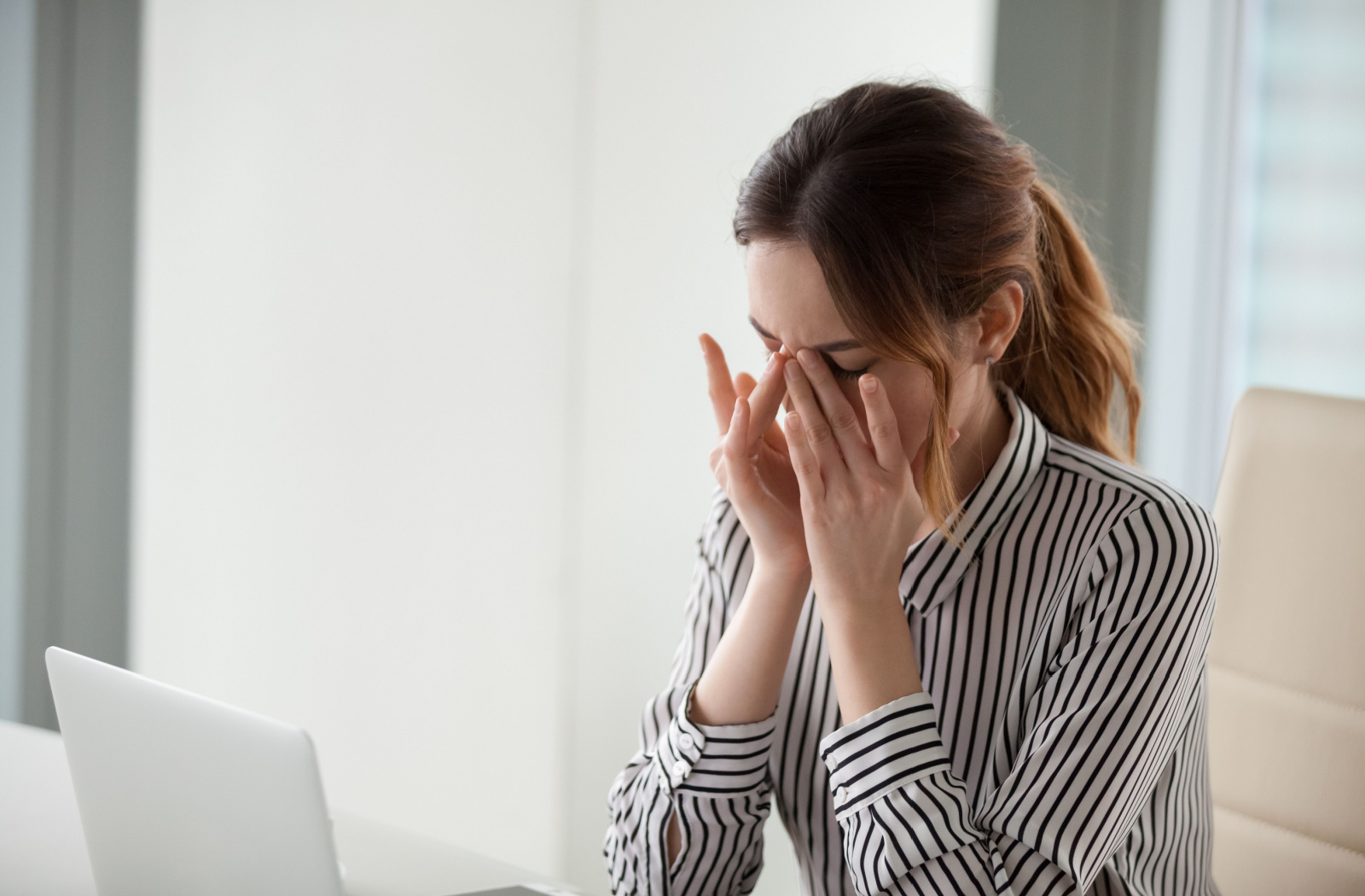 A young woman sitting at a desk with her laptop as she rubs her eyes
