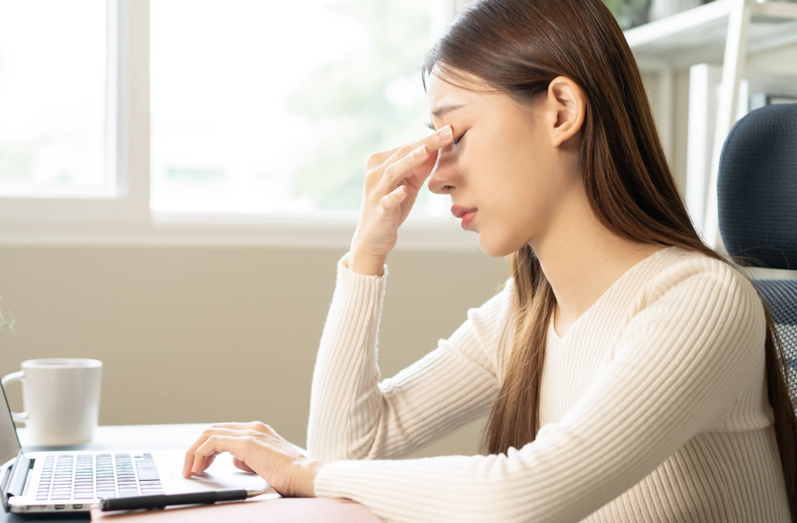 A young woman in a sweater squeezing the bridge of her nose with her right hand while working on her laptop.
