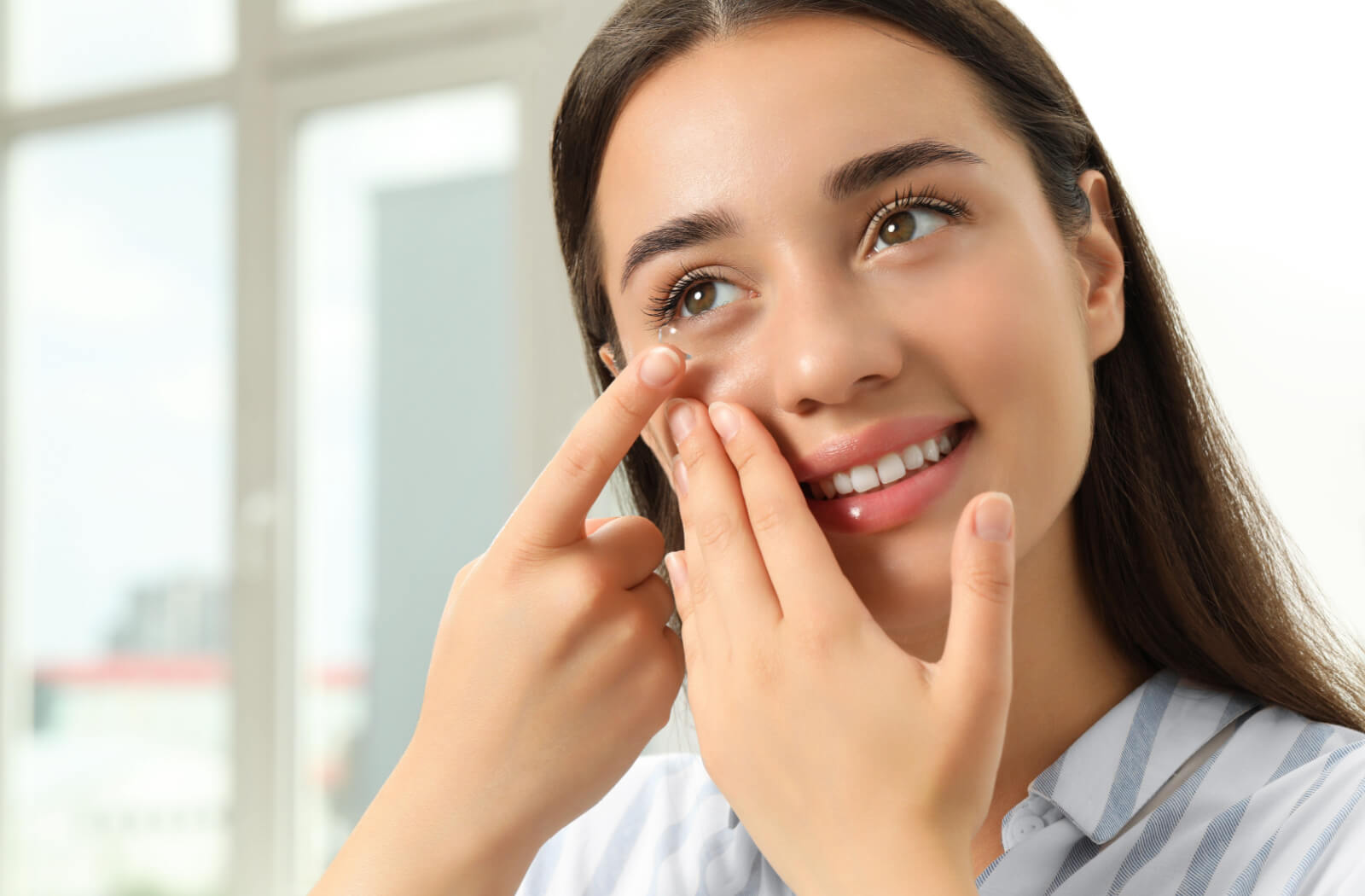A woman using her left fingers to pull her eyelid down while she puts a contact lens on her right eye with her right hand