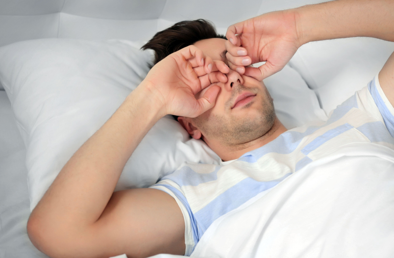 A young man lying in bed rubbing his eyes with his hands.