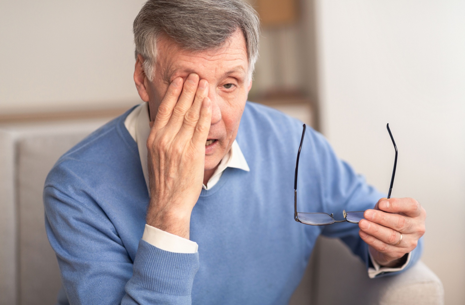 An older adult man sitting on a couch and holding his glasses in his left hand as he rubs his right eye with his other hand