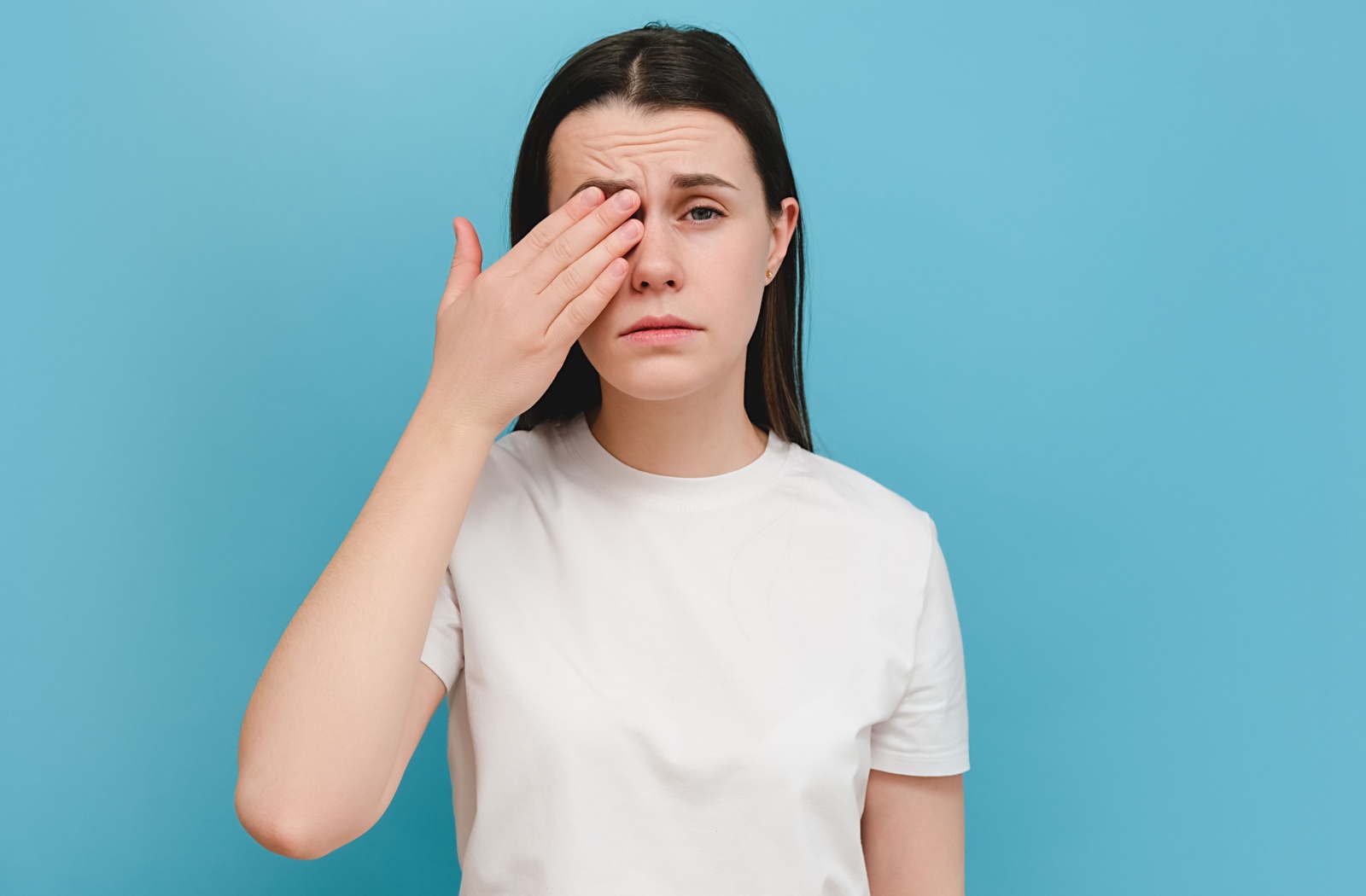 A young person, against a blue background, covers their left eye with their hand.