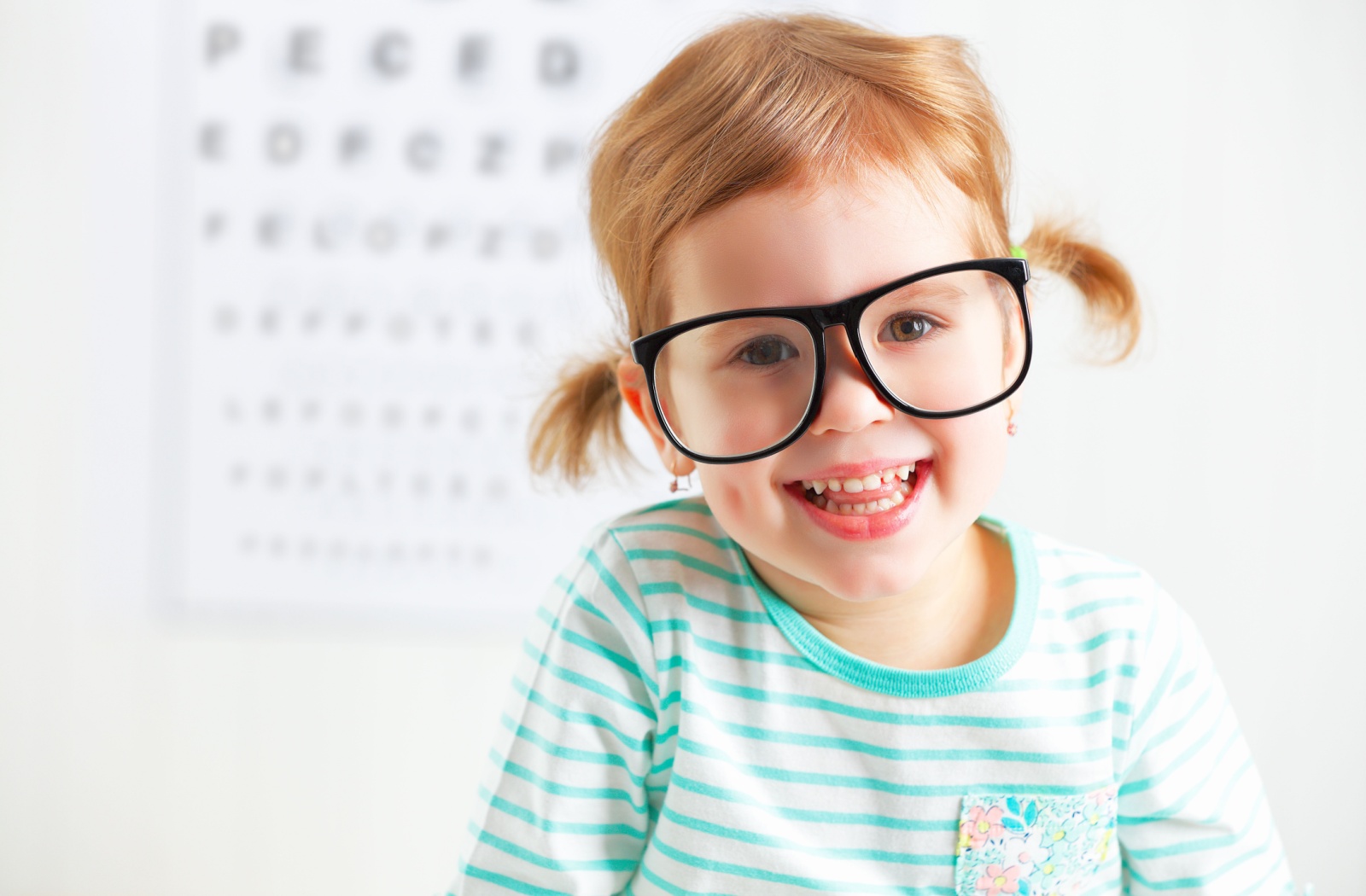 Young child wearing glasses at an eye exam with vision chart in background.